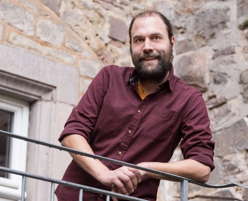 Mitarbeiterporträt eines Mannes mit Bart vor Steinwand