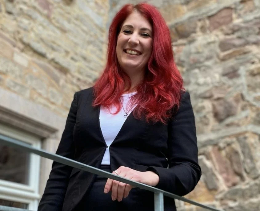 Mitarbeiterporträt einer Frau mit roten Haaren vor Steinwand